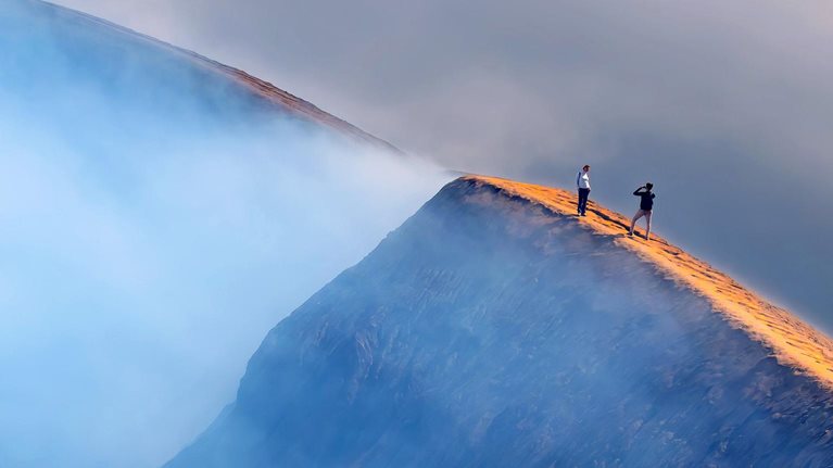 Aerial view of a mountain under clouds. Two walkers take photographs.