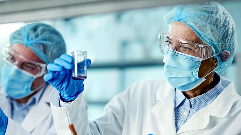 Two scientists in a laboratory are wearing protective gear including lab coats gloves and face masks One scientist carefully examines a liquid in a beaker while the other holds test tubes containing samples.