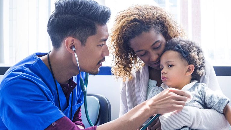 A young doctor in a blue scrub is using a stethoscope to examine a toddler who is sitting on their mother's lap.