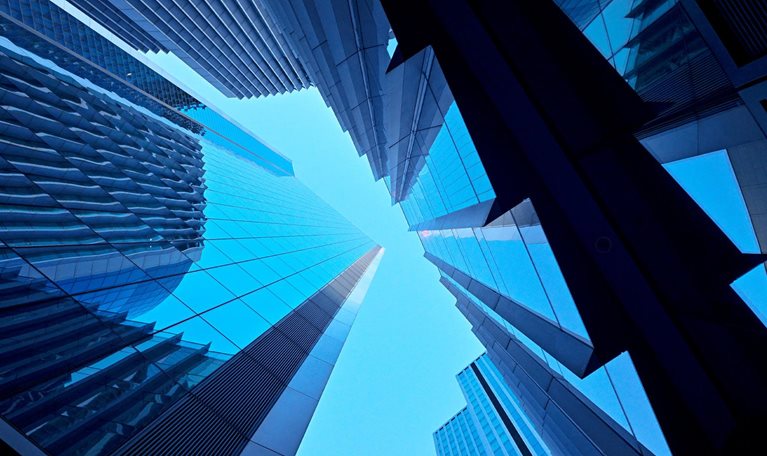 A dramatic upward view of modern glass skyscrapers converging toward the sky. The sharp lines and reflective surfaces create a sleek, futuristic feel.