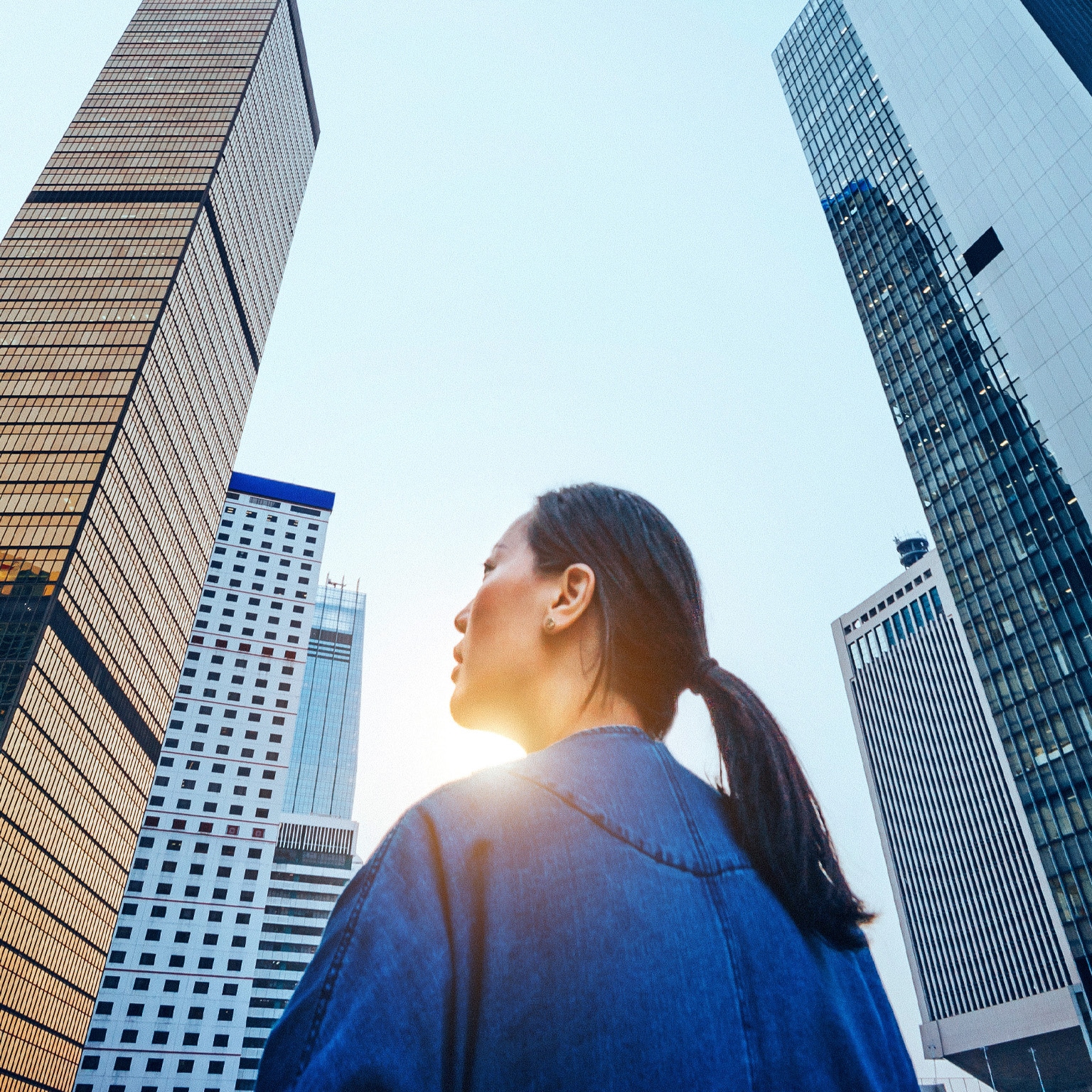 A woman stands with her back to the camera, gazing upward at a cluster of towering skyscrapers that dominate the frame. 