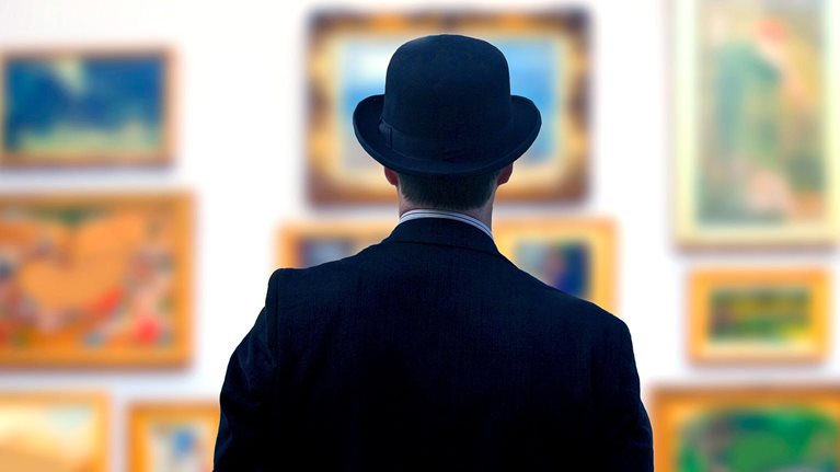 A man in a black suit and bowler hat looks at art in a gallery.
