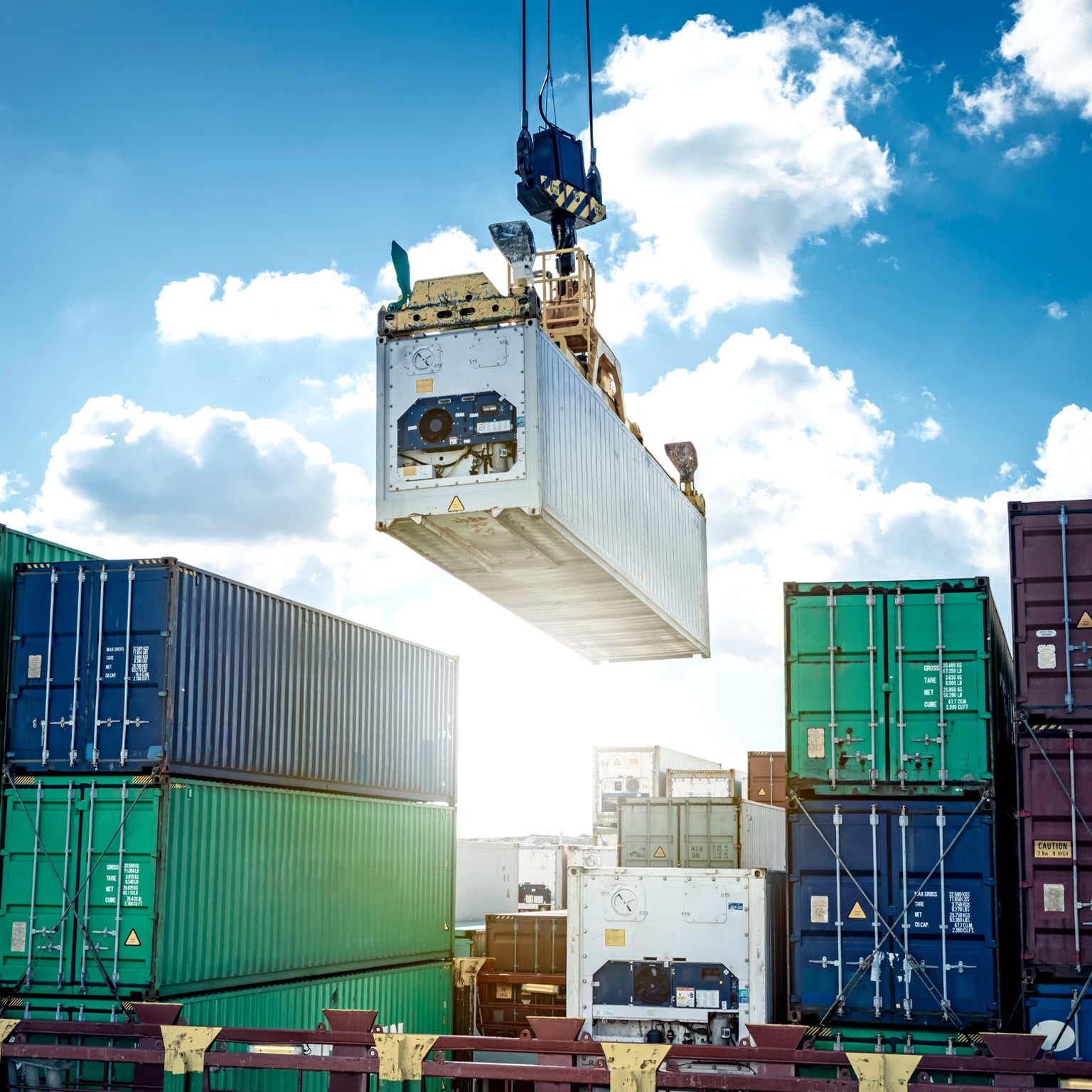 A large crane lifting a shipping container high above stacks of containers at a busy port. The bright sky and orderly rows of containers emphasize large-scale cargo handling and global trade operations.