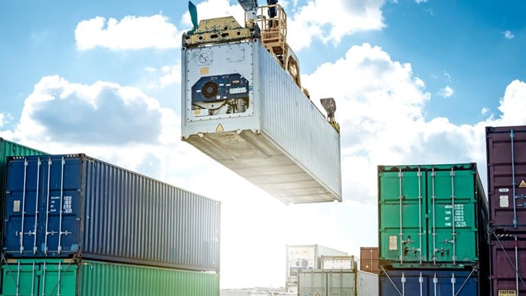 A large crane lifting a shipping container high above stacks of containers at a busy port. The bright sky and orderly rows of containers emphasize large-scale cargo handling and global trade operations.