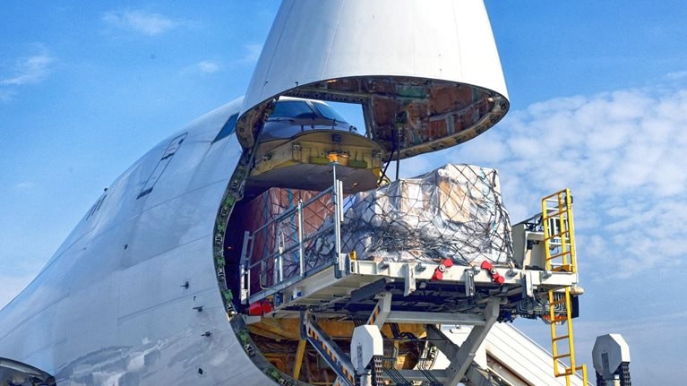 A cargo plane has its front end open as a sizable pallet is being loaded onto it.