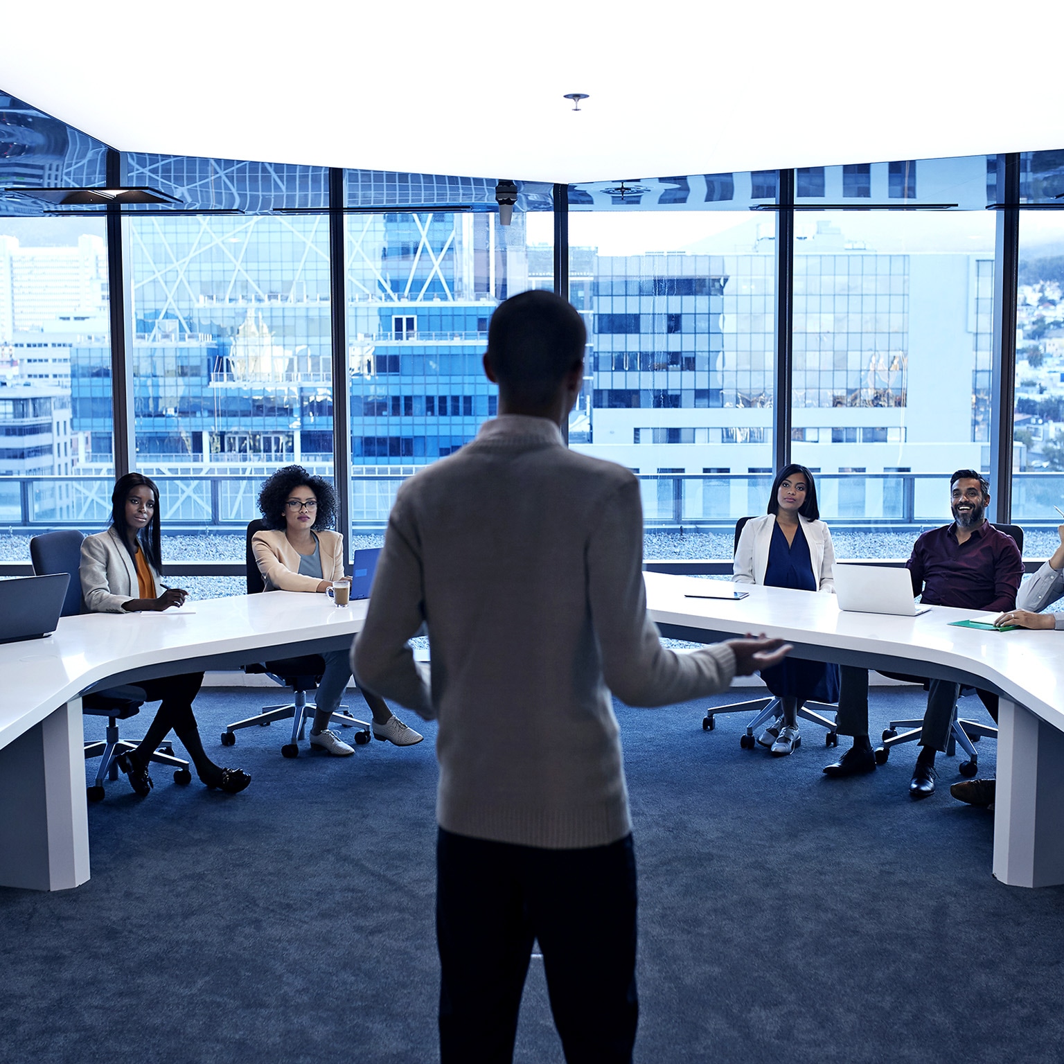 Image of a man standing at the head of a curved white table in a modern office, addressing diverse professionals in a meeting.