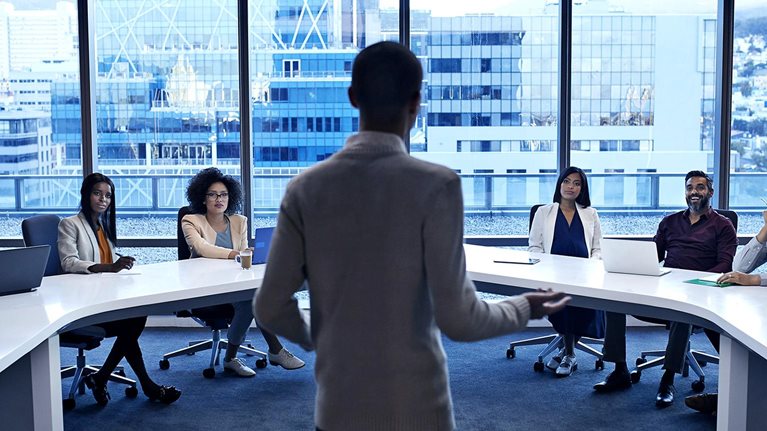 Image of a man standing at the head of a curved white table in a modern office, addressing diverse professionals in a meeting.