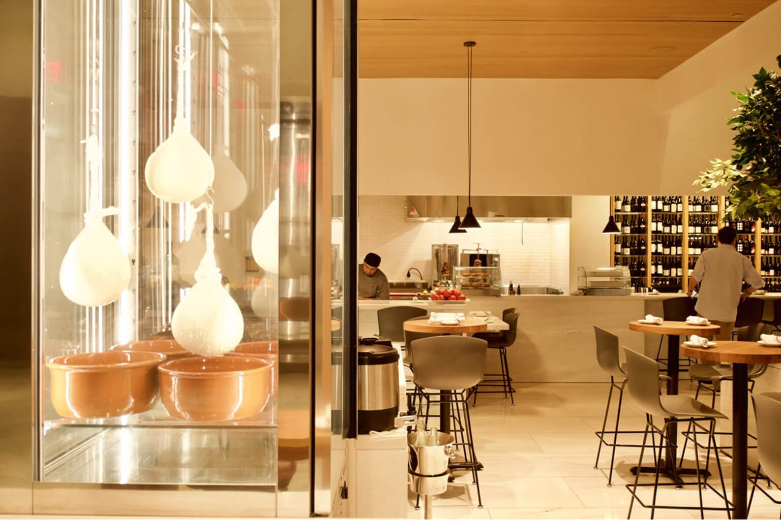 The image displays the interior of the Milos Wine Bar, which is part of the estiatorio Milos restaurant at Hudson Yards in New York City. The space features a display case with hanging cheese and an open kitchen concept.