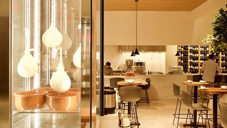 The image displays the interior of the Milos Wine Bar, which is part of the estiatorio Milos restaurant at Hudson Yards in New York City. The space features a display case with hanging cheese and an open kitchen concept.