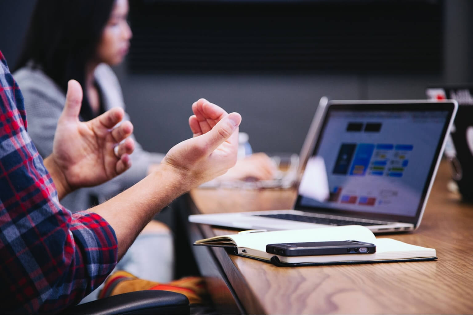 The image depicts a business meeting or presentation in progress. The scene shows individuals in a professional setting collaborating or discussing information. A person in a plaid shirt is actively gesturing while another person is seated in the background.