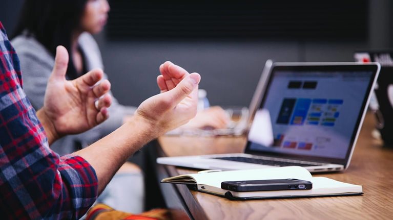 The image depicts a business meeting or presentation in progress. The scene shows individuals in a professional setting collaborating or discussing information. A person in a plaid shirt is actively gesturing while another person is seated in the background.