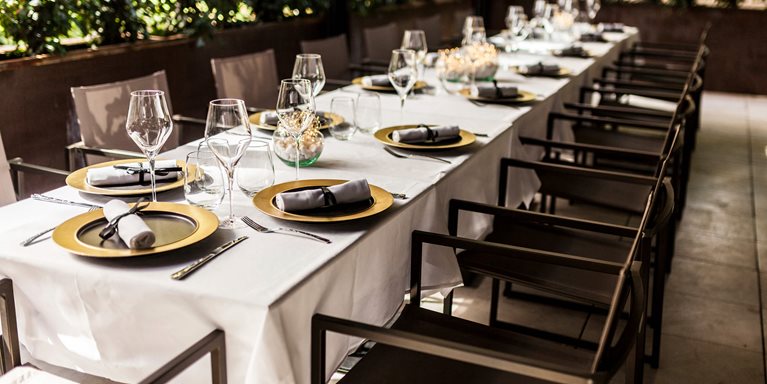 The image features an elegant restaurant terrace set for a formal breakfast, featuring a long table dressed in a crisp white cloth. Each place setting includes striking gold charger plates, gray napkins, modern stemware, and polished silverware, ready to accommodate a group for a special occasion or private event.