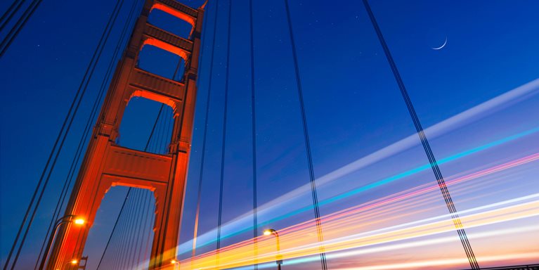 Golden Gate Bridge in San Francisco, California. The photo captures the glowing orange hue of one of the bridge's main steel towers against a deep blue dusk sky, complemented by the glow of a slender crescent moon.