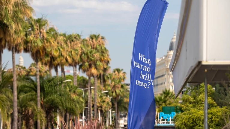 This image was captured during the Cannes Lions International Festival of Creativity. It features a large, blue feather flag prominently displaying the text What's your next brilliant move?