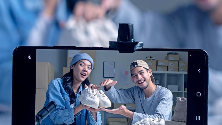 Two people hold up a pair of new sneakers toward a mobile-phone camera, with a microphone in the foreground indicating audio in addition to video recording