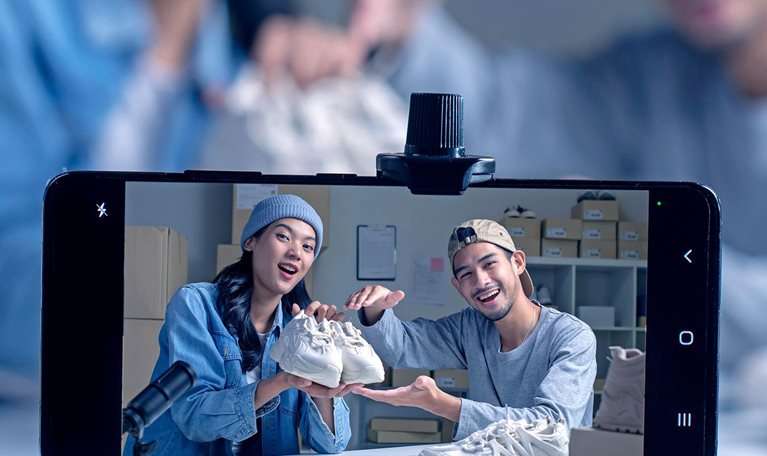 Two people hold up a pair of new sneakers toward a mobile-phone camera, with a microphone in the foreground indicating audio in addition to video recording