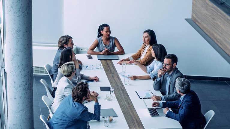 In a modern office setting with bright lighting and large windows, a diverse group of professionals gathers around a long conference table, seemingly engaged in a business meeting.