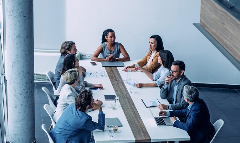 In a modern office setting with bright lighting and large windows, a diverse group of professionals gathers around a long conference table, seemingly engaged in a business meeting.