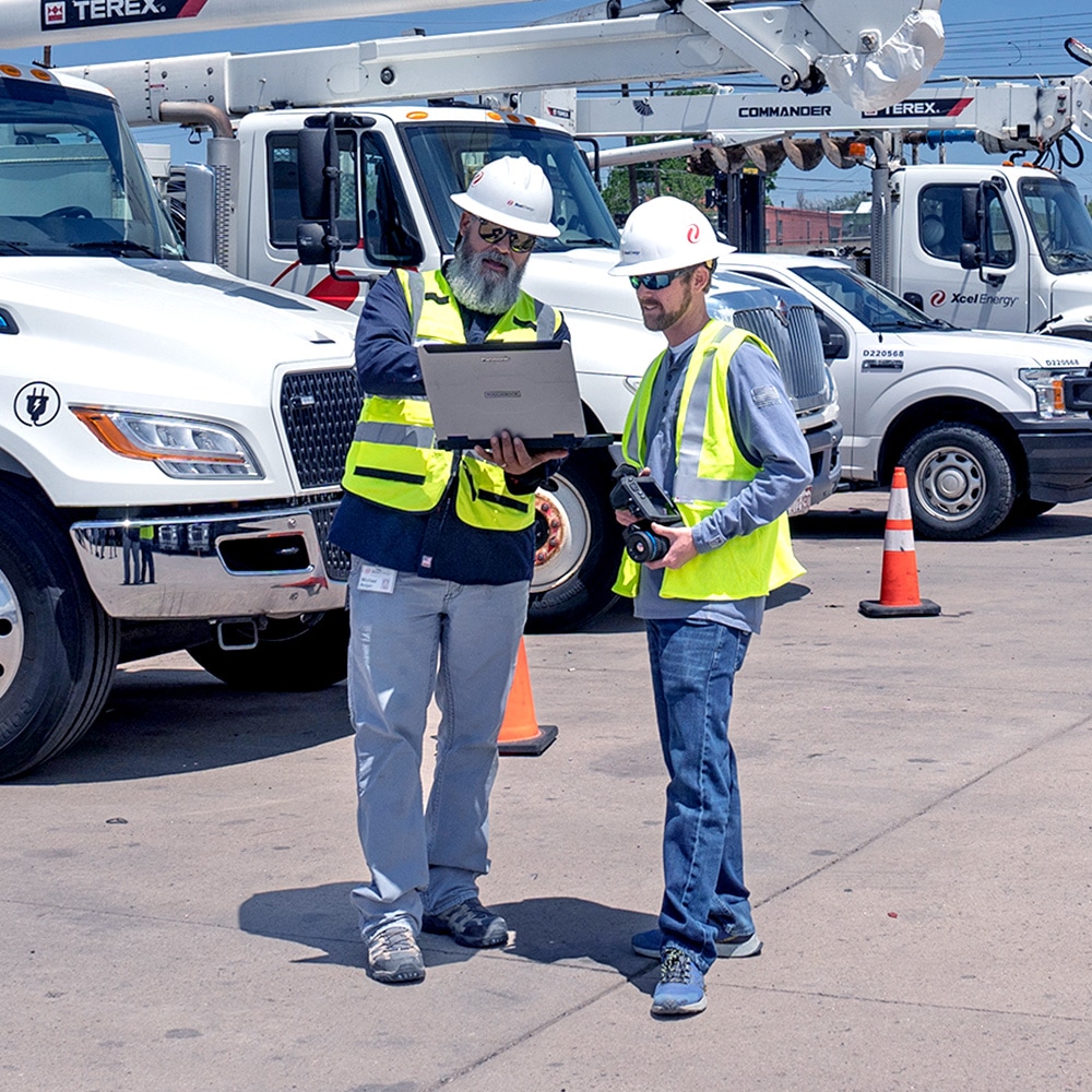 Two Xcel workers at a job site reviewing plans on a laptop