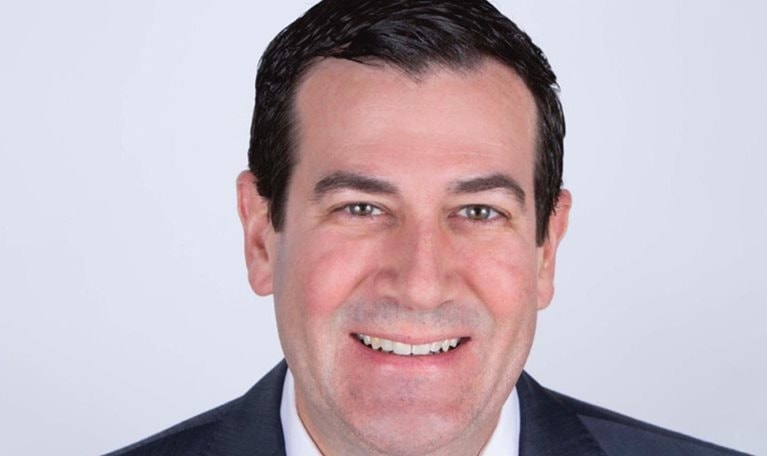 Portrait of Mark Birkhead with a light smile, wearing a charcoal suit and a maroon tie.