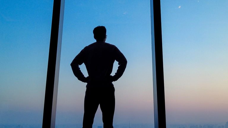 Young man staring out floor to ceiling office window looking over city skyline