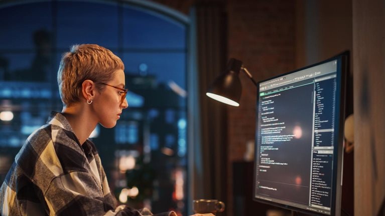 Young Woman Writing Code on Desktop Computer in Stylish Loft Apartment in the Evening. Creative Female Wearing Cozy Clothes, Working from Home on Software Development. Urban City View from Big Window. - stock photo