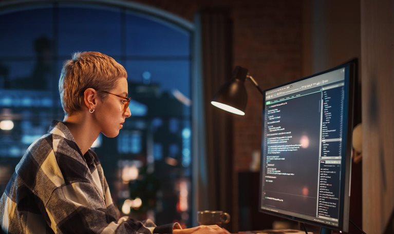 Young Woman Writing Code on Desktop Computer in Stylish Loft Apartment in the Evening. Creative Female Wearing Cozy Clothes, Working from Home on Software Development. Urban City View from Big Window. - stock photo