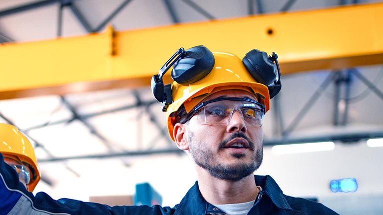 A focused male warehouse worker in safety gear inspects machinery with another worker, ensuring industry standards and safety protocols in a well-lit factory setting.