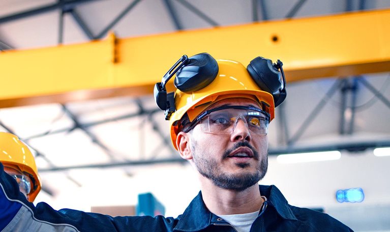 A focused male warehouse worker in safety gear inspects machinery with another worker, ensuring industry standards and safety protocols in a well-lit factory setting.