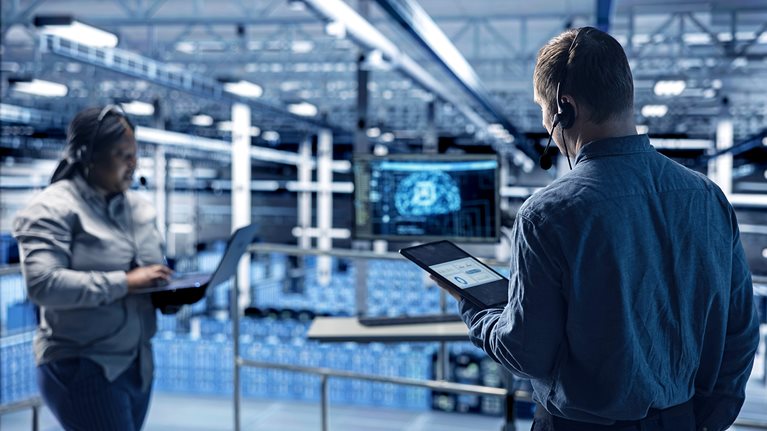 Two IT professionals work with laptops and tablets in a modern server room, managing data and technology infrastructure