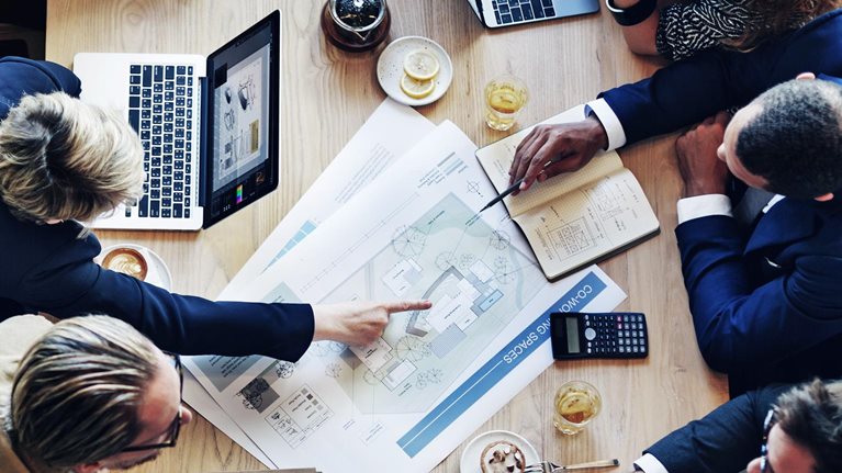 From a bird's-eye view, a diverse group of business professionals is gathered around a table covered with laptops, tablets, design plans, and financial reports The team is engaged in a spirited conversation about a project.