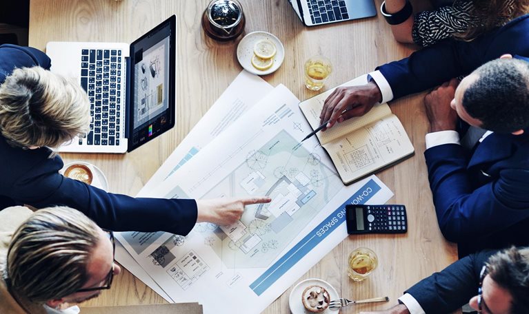 From a bird's-eye view, a diverse group of business professionals is gathered around a table covered with laptops, tablets, design plans, and financial reports The team is engaged in a spirited conversation about a project.