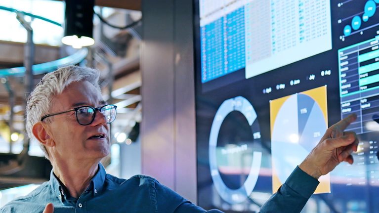 A man gesturing towards a large screen displaying graphs and data as he leads a presentation.