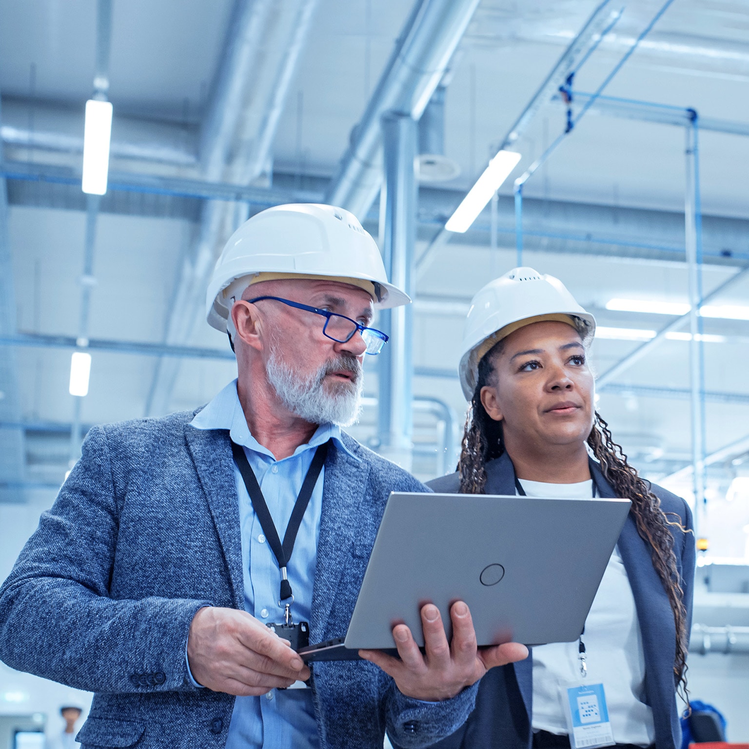 A male and female engineer in hard hats walking on factory floor and discussing the facility's machinery. 