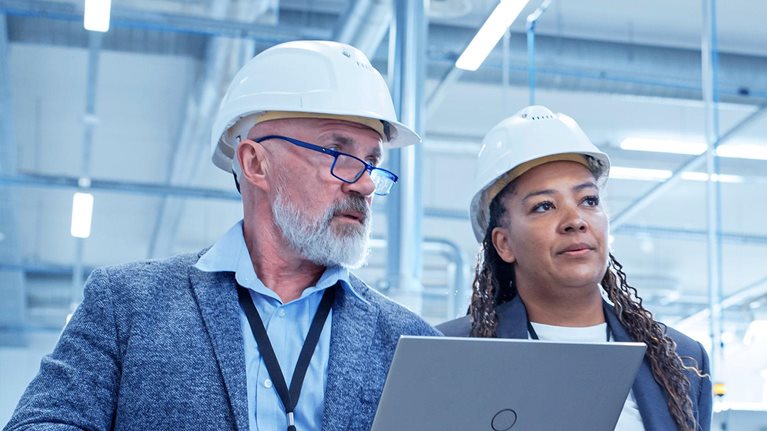 A male and female engineer in hard hats walking on factory floor and discussing the facility's machinery.