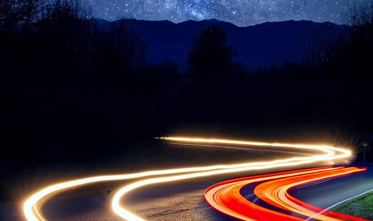 car headlight trails driving on a road through a forest on a starry night with the milky way and mountains in the background