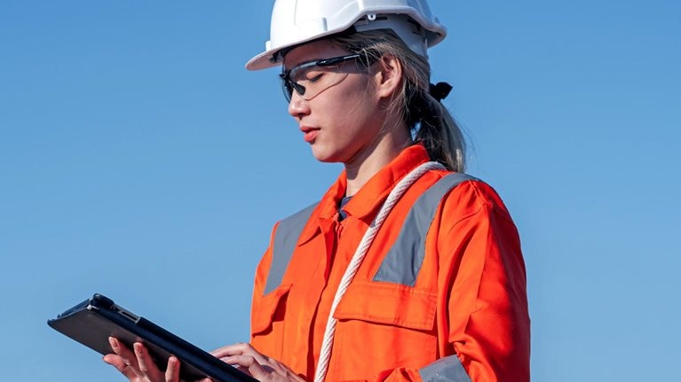 Image of a woman at work using a tablet computer, wearing a hard hat, safety glasses, and a bright colored safety shirt.