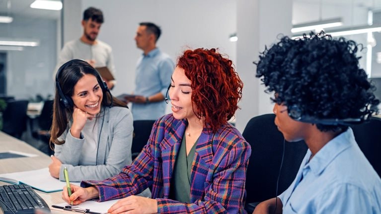 A technical support manager at a call center sits in between two colleagues as she instructs them on how to assist customers.
