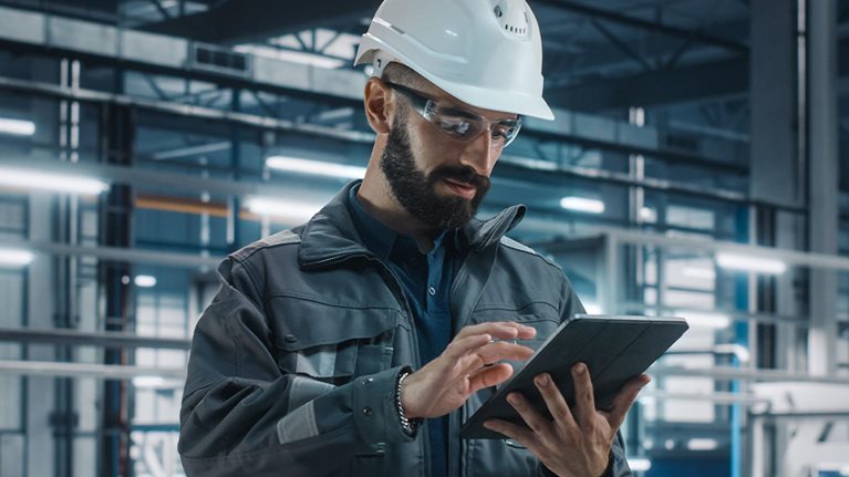 Portrait of Automotive Industry Engineer in Safety Glasses and Uniform Using Laptop at Car Factory Facility. Professional Assembly Plant Specialist Working on Manufacturing Modern Electric Vehicles.