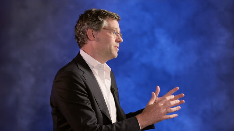 Erikhans Kok wearing glasses, a dark suit jacket, and a white shirt gestures with both hands while facing to the right against a blue background.