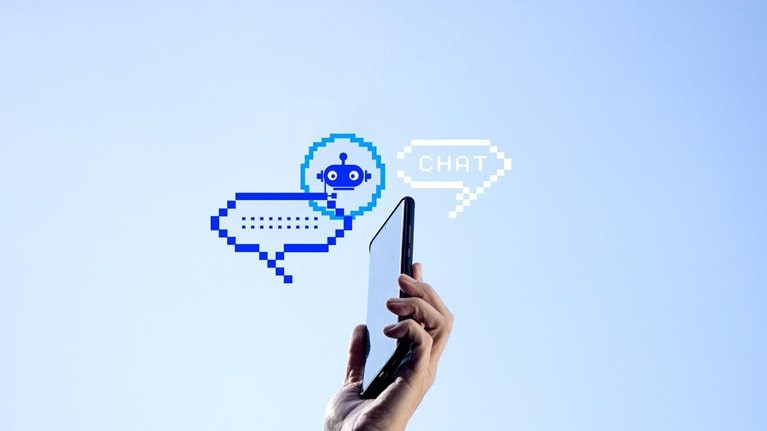 A hand holding a mobile phone with two pixelated chat bubbles and an AI bot icon against a clear blue sky.