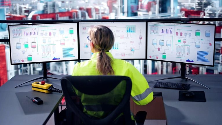 Image of an engineer operating an industrial data automation system using multiple computer screens with a factory in the background.