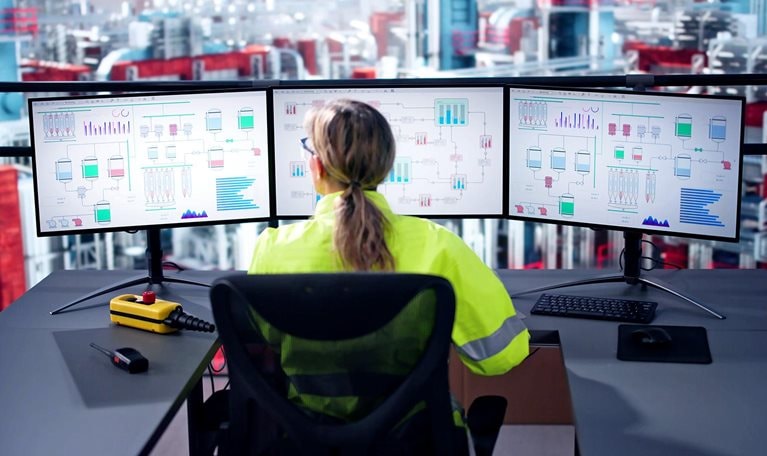 Image of an engineer operating an industrial data automation system using multiple computer screens with a factory in the background.