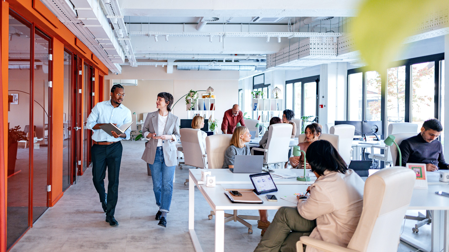 A bustling, modern office space with a diverse group of employees engaged in various work activities, some collaborating in groups, while others work individually at their desks. Two colleagues are seen walking and talking in a hallway, showcasing a dynamic and collaborative work environment. 