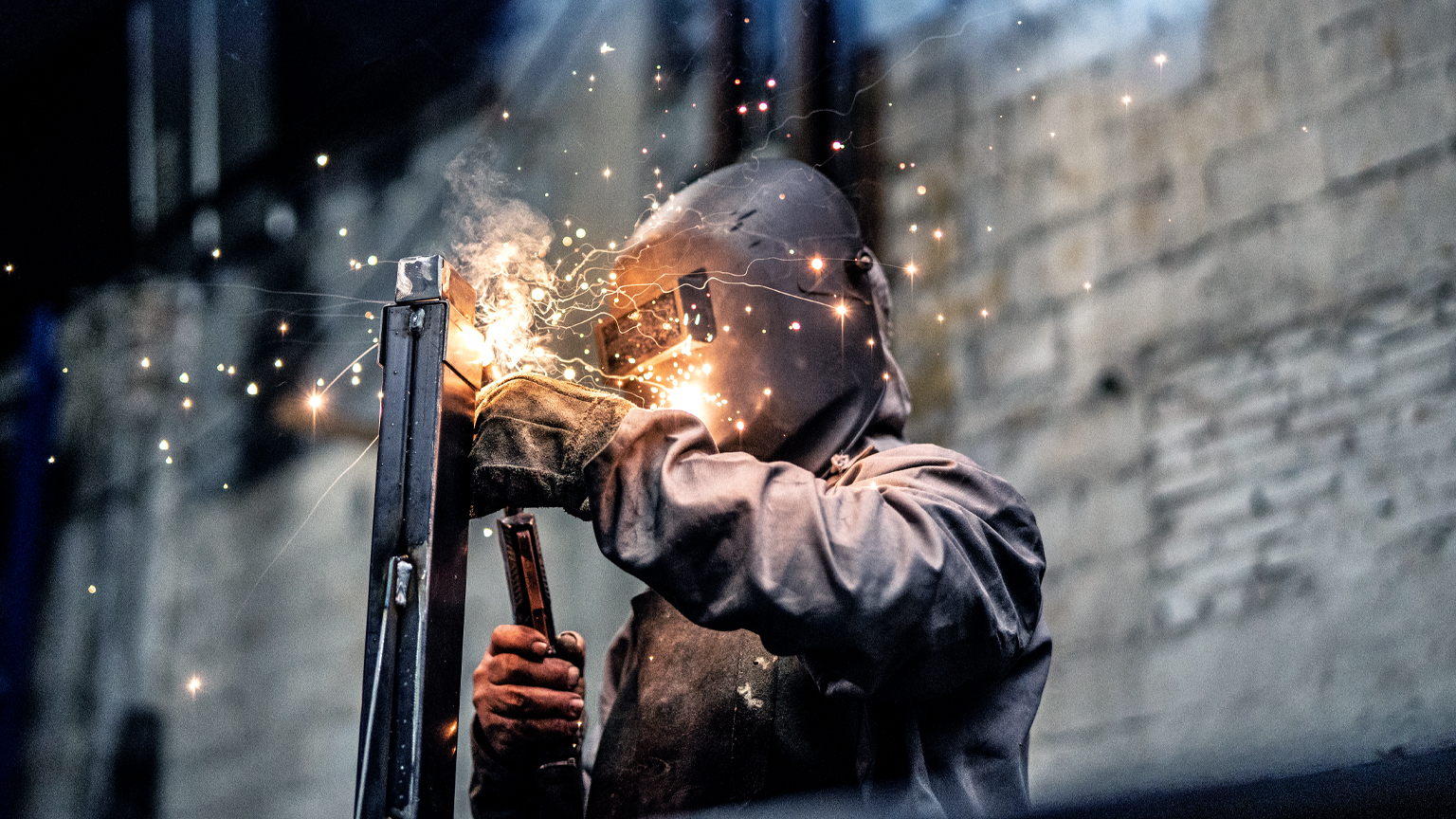 An individual clad in protective welding attire, working on a steel component while sparks scatter.