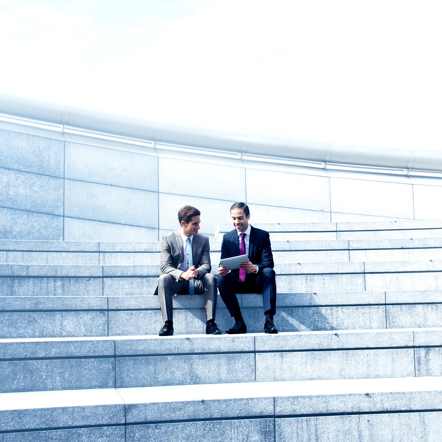 A manager giving feedback to an employee. They are sitting on a set of stairs while looking at a tablet.