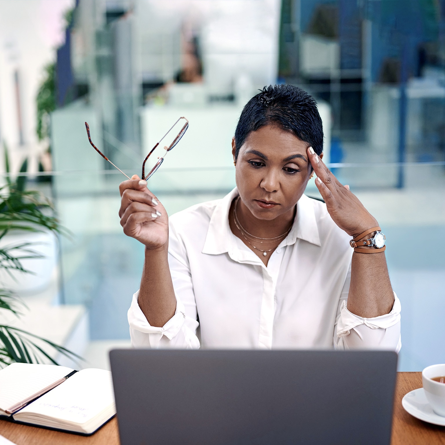 Woman looking at a laptop holding her glasses in one hand and pressing the fingers of her other hand into her temple.