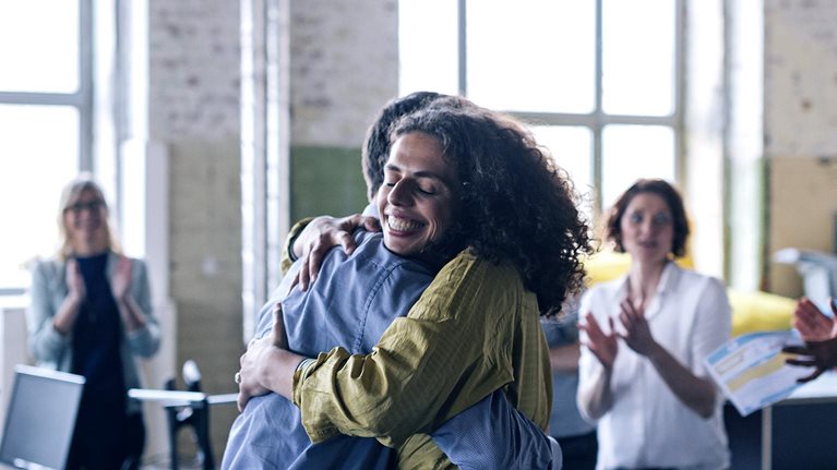 Image of people celebrating in an office with a woman smiling and being hugged by her coworker.