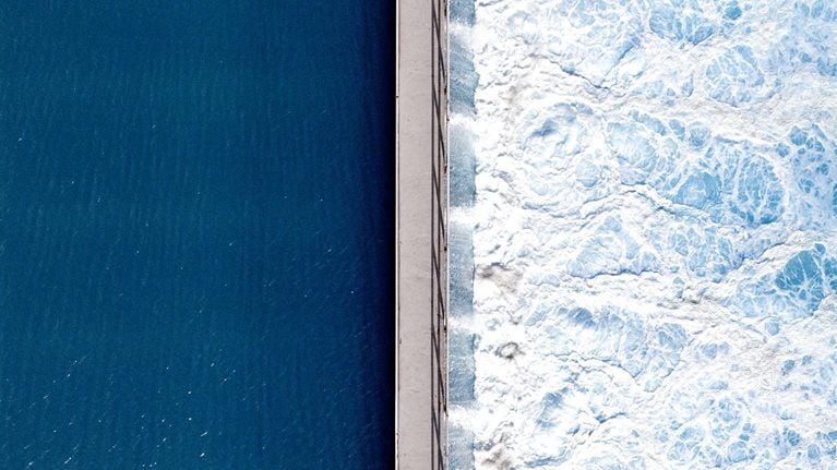 An overhead shot captures the stark contrast between deep, tranquil blue water on one side and churning, white-foamed turquoise waves on the other, bisected by a narrow concrete pier. The smooth, dark surface on the left stands in dramatic opposition to the textured, frothy turbulence crashing against the right side of the man-made barrier.