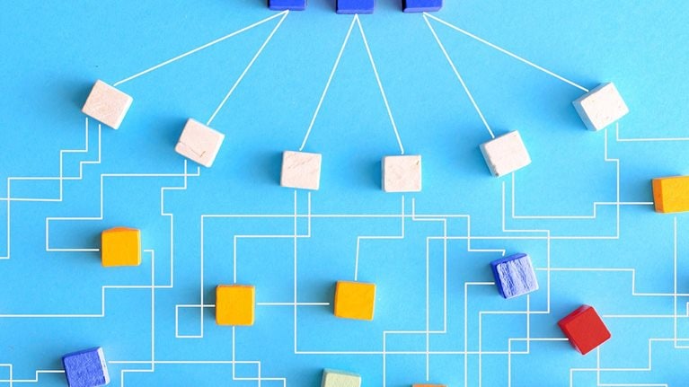 An organization chart constructed from wooden blocks that are linked together with delicate white lines. At the uppermost level of the chart, there are three blue blocks that connect to six white blocks. These white blocks then link outwards to form a larger network of blocks in varying shades of orange, red, and blue.